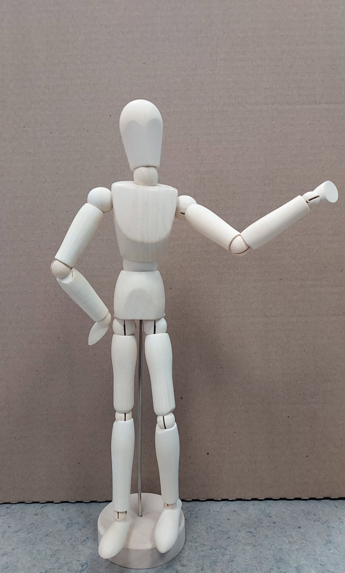 A wooden artist mannequin stands upright on a round base, with one arm bent at the hip and the other arm raised and extended outward against a plain cardboard background.