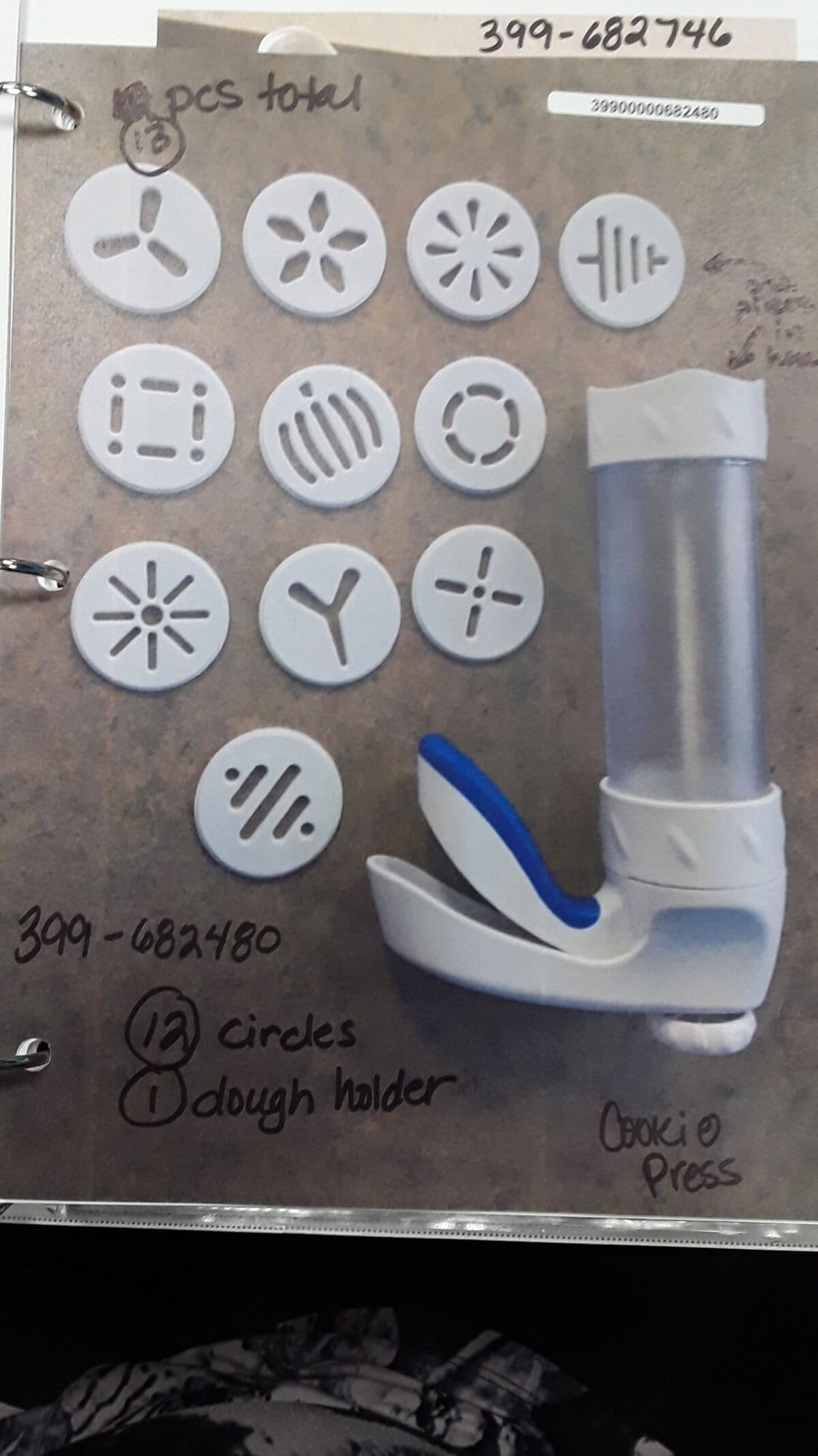 A cookie press with a blue handle is shown next to 13 circular dough discs, each with different cut-out patterns, arranged on a tabletop. Handwritten notes label the items and provide product numbers.