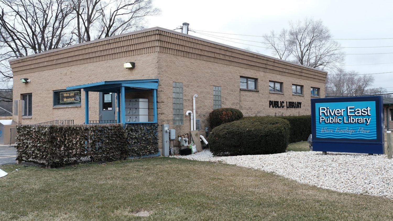 A small, tan brick building with a blue sign reading River East Public Library in front, surrounded by grass, shrubs, and bare trees on a cloudy day.