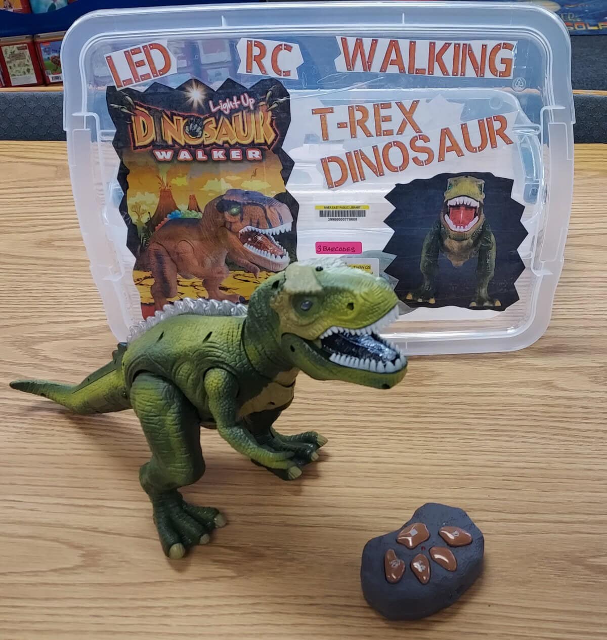A green toy T-Rex dinosaur stands on a wooden surface in front of its plastic storage box, which is labeled LED RC WALKING T-REX DINOSAUR. A brown toy remote with four buttons is beside the dinosaur.