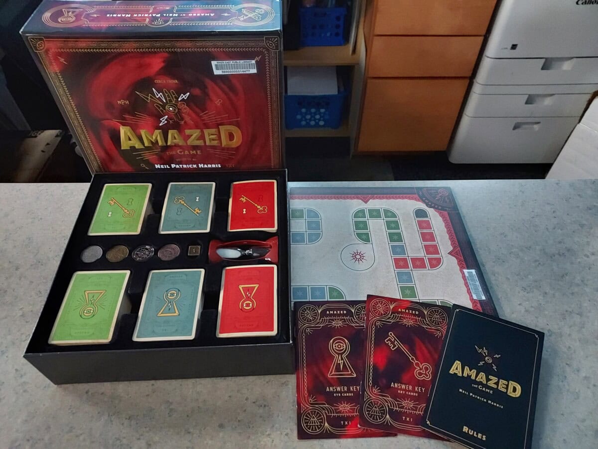 A board game called AMAZED is displayed on a table. The open box reveals cards, coins, a spinner, and a game board with colored paths. The rules and answer key booklets are also visible.