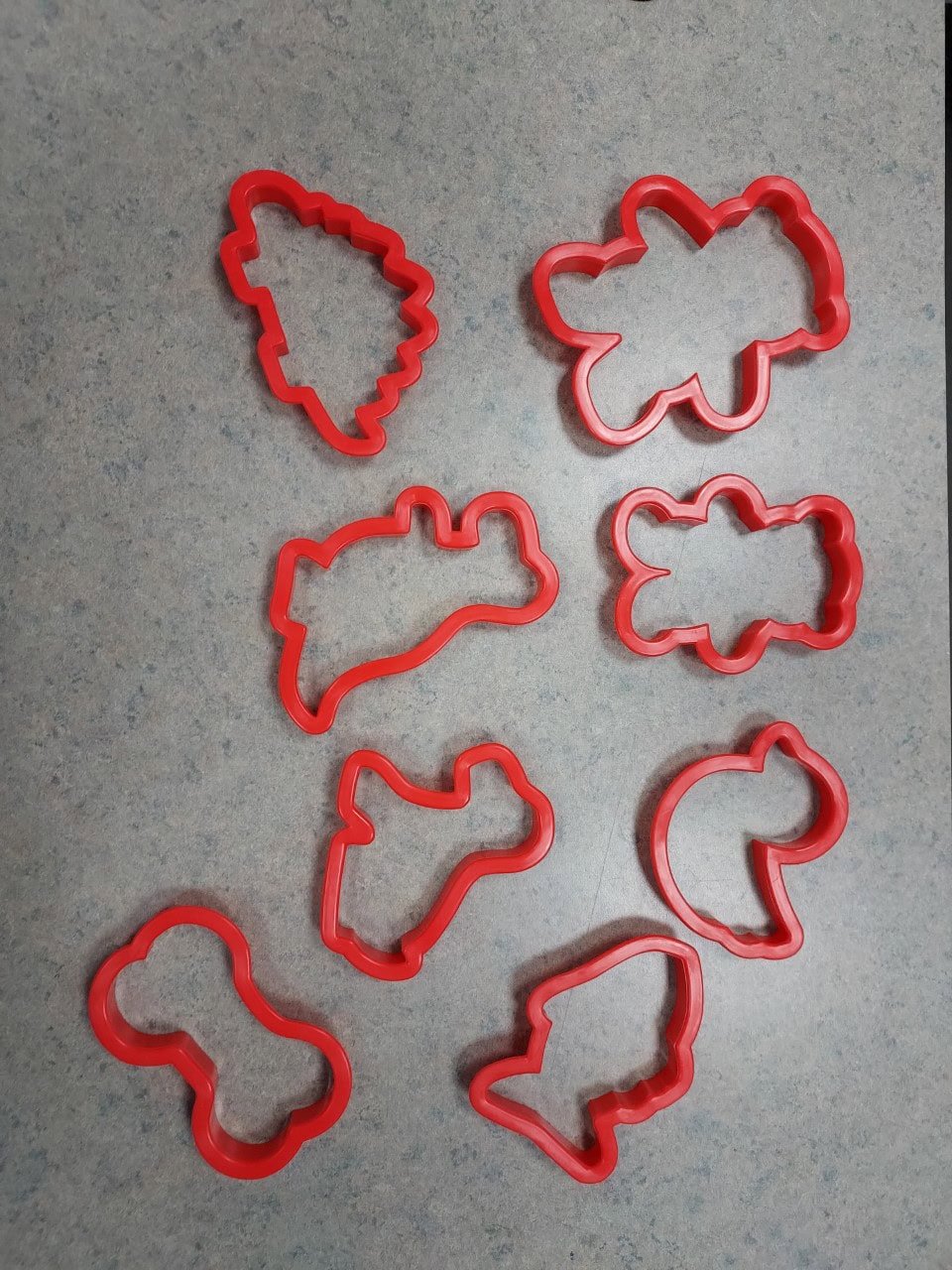 Nine red plastic cookie cutters of various shapes, including a Christmas tree, bone, dog, cat, apple, fish, and other animals, are arranged on a gray speckled surface.