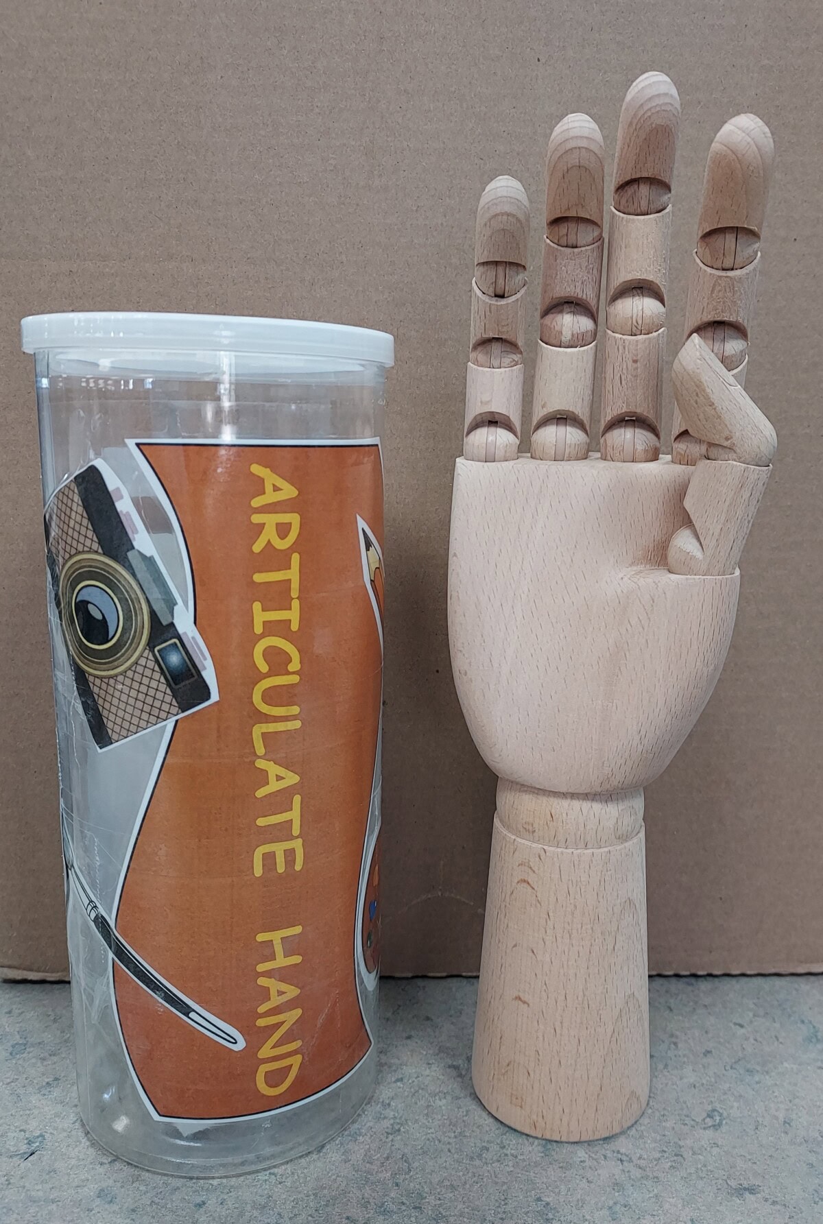 A wooden articulated hand model stands upright next to a clear plastic container labeled ARTICULATE HAND with illustrated art supplies on the label. The background is plain cardboard.