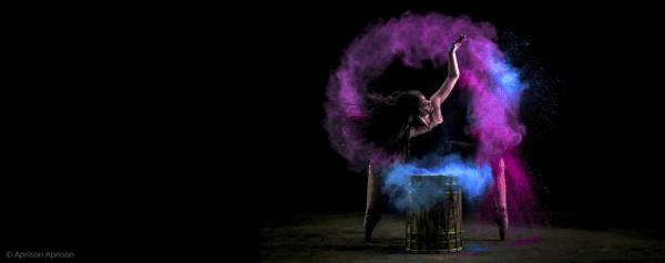 A dancer bends backward dramatically behind a drum, tossing vibrant clouds of pink and blue powder into the air against a black background.