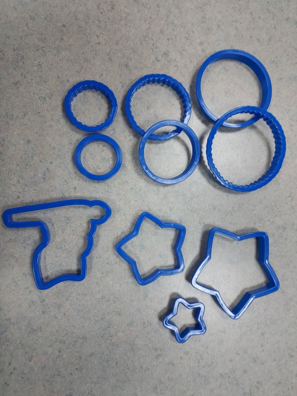 A set of blue plastic cookie cutters on a gray surface, including round cutters with smooth and scalloped edges, and cutters shaped like stars and a pistol.