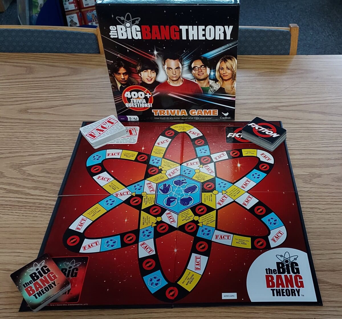 A Big Bang Theory trivia board game is set up on a wooden table. The board has an atom design with cards, tokens, and the game’s box featuring main cast members in the background.