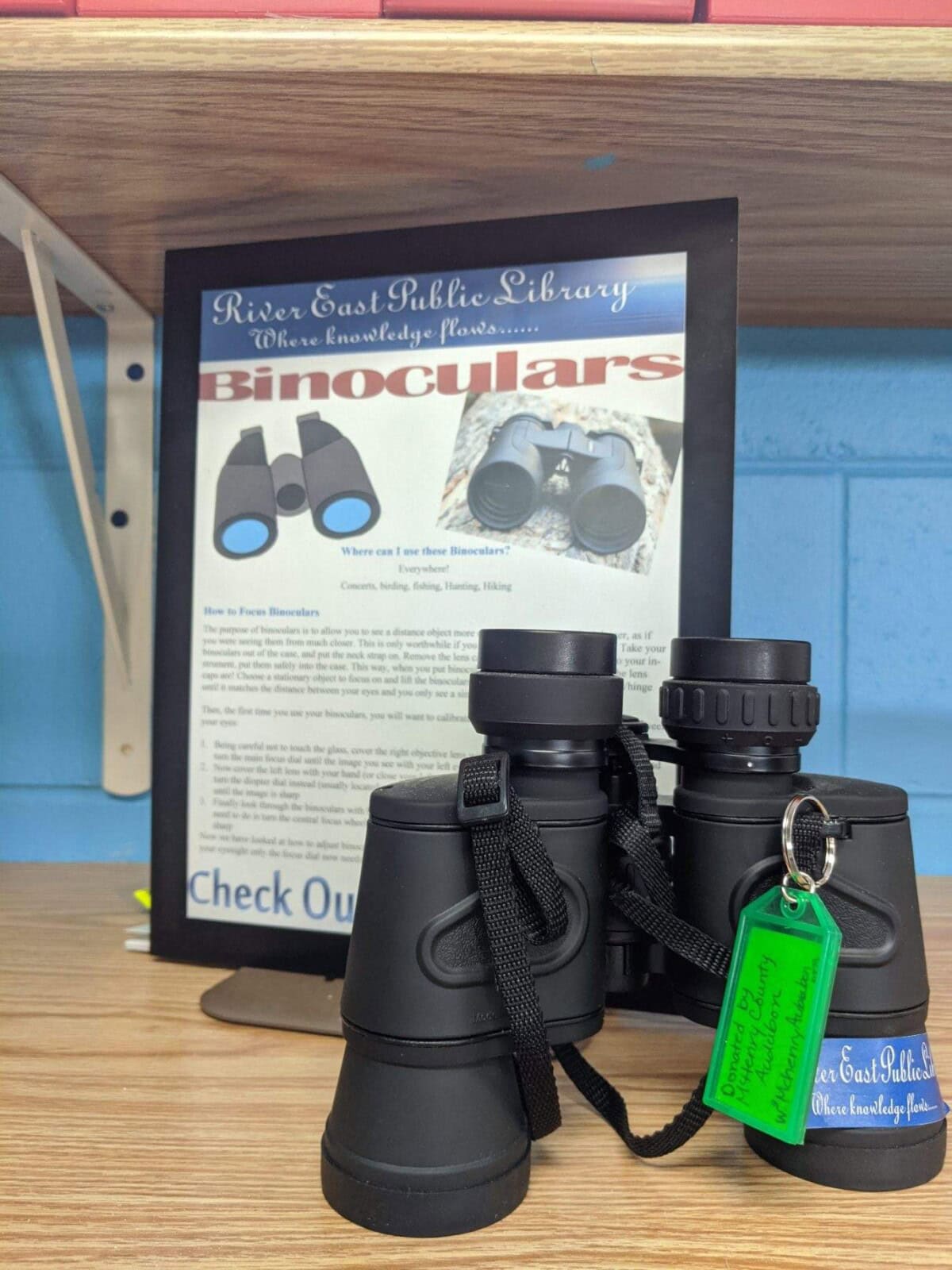 Two black binoculars with straps rest on a wooden shelf in front of a sign about checking out binoculars from the River East Public Library. The right binoculars have a green tag attached to them.
