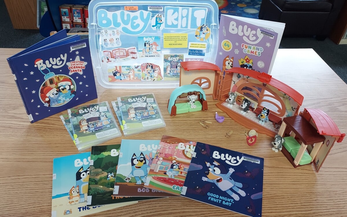 A display of Bluey-themed items on a table, including books, DVDs, playsets with figures, a Bluey Kit box, and activity materials, featuring colorful illustrations from the animated series Bluey.