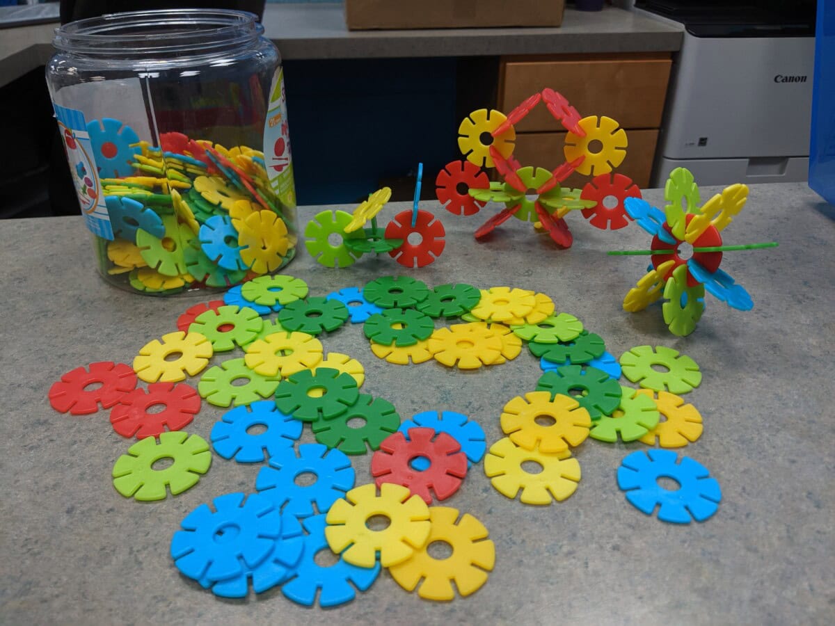 Colorful plastic interlocking discs, mostly blue, green, yellow, and red, are scattered on a tabletop. Some are built into 3D shapes, with a large jar of loose pieces in the background.