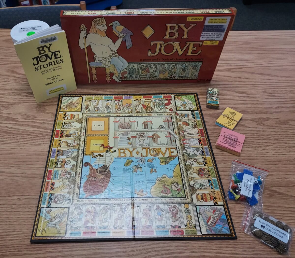 A board game called By Jove displayed on a wooden table with its box, game board showing illustrations, rulebooks, cards, and game pieces in a plastic bag. The game has a mythology theme.