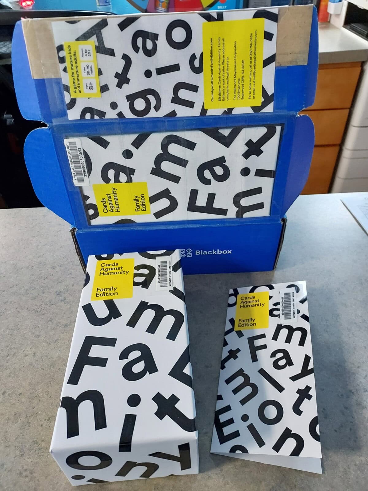 Two boxes and two card packs of Cards Against Humanity: Family Edition with bold black and white text on the packaging, displayed on a countertop, with some kitchen items blurred in the background.