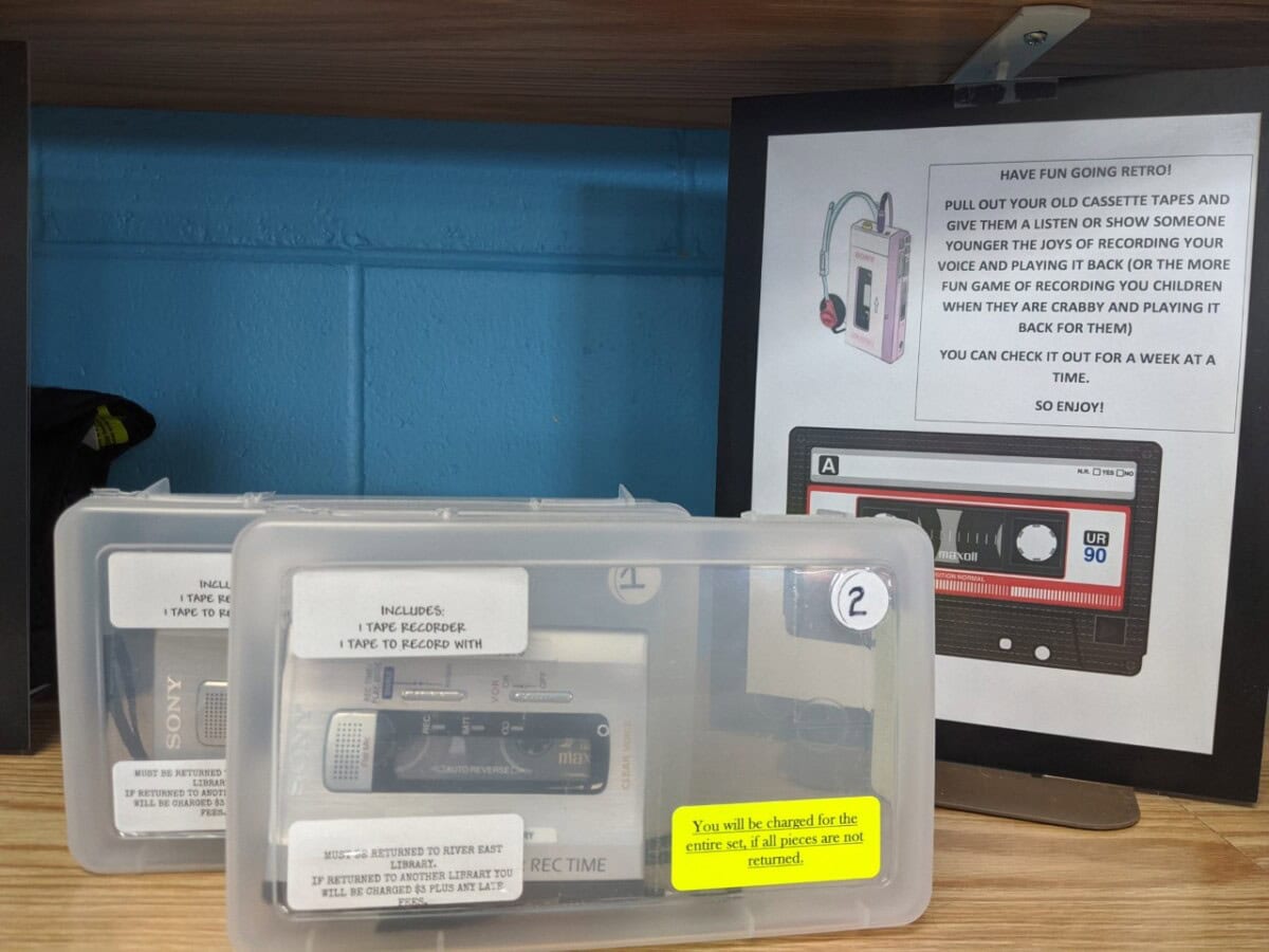 Two plastic kits containing tape recorders and tapes sit on a wooden shelf. Behind them, a sign encourages people to borrow a cassette player for a week to revisit old tapes or record memories.