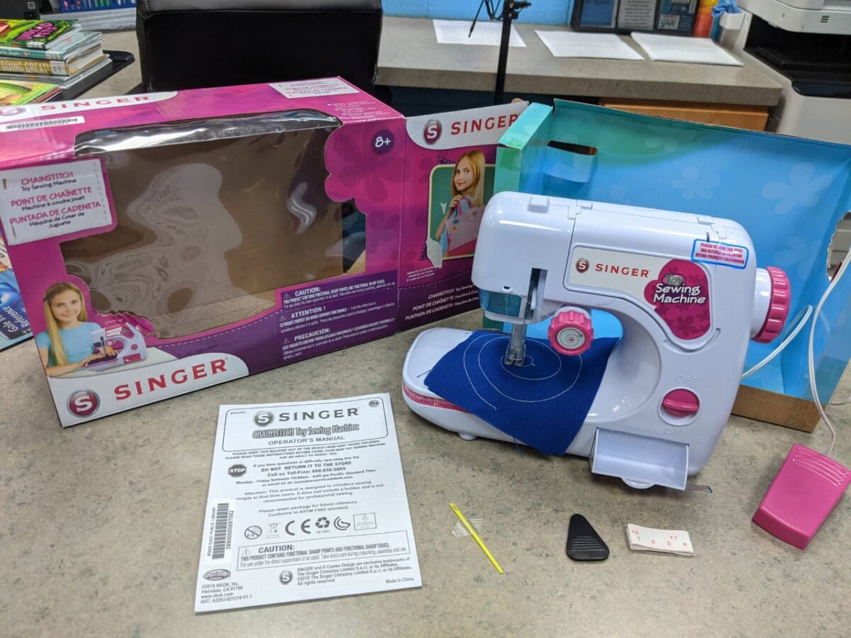 A small Singer toy sewing machine is on a table next to its pink box, instruction sheet, and sewing accessories. The machine is set up with blue fabric under the presser foot.