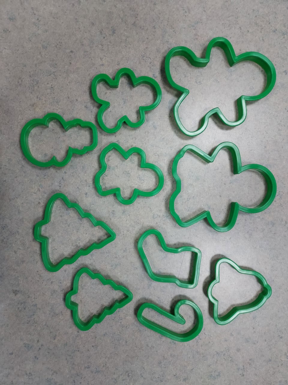 Green plastic cookie cutters in various holiday shapes, including gingerbread people, Christmas trees, a stocking, a candy cane, and clouds, are arranged on a gray countertop.