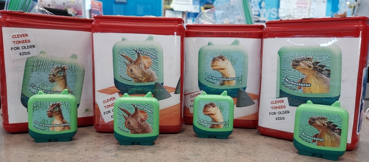 Four red and white containers with dinosaur images and Clever Tonies for Older Kids text behind five small green dinosaur-themed toys on a countertop, each labeled with a different dinosaur name.
