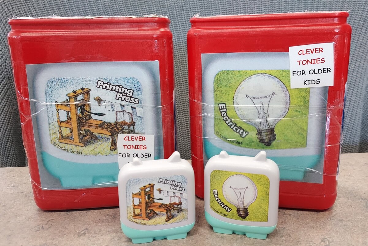 Two red plastic containers and two white figurines feature illustrations of a printing press and a lightbulb labeled Printing Press and Electricity. The labels read CLEVER TONIES FOR OLDER KIDS.