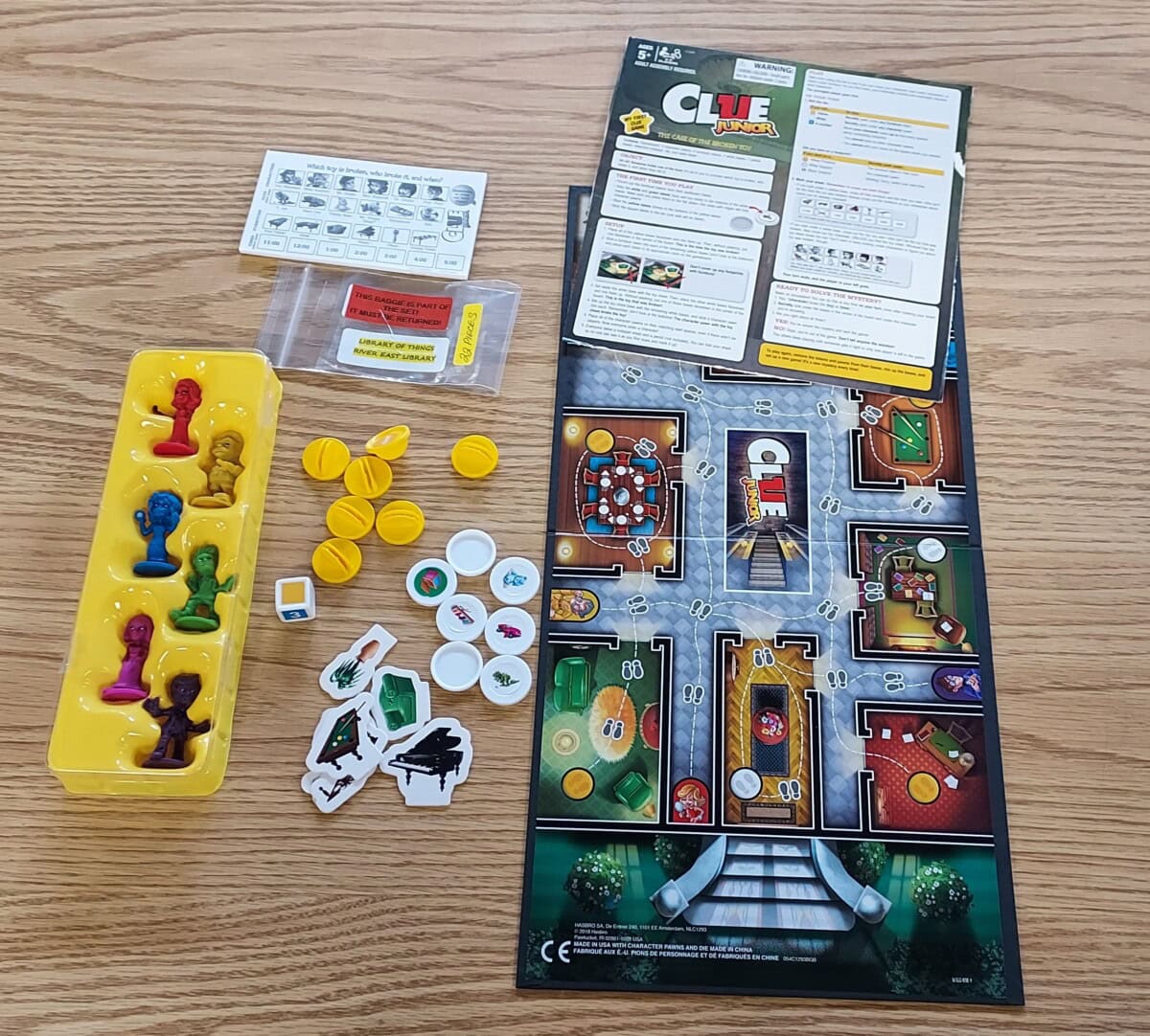 Top-down view of the Clue Junior board game set up on a wooden table, showing the board, cards, yellow tokens, plastic figures in various colors, clue tokens, and instruction sheets.