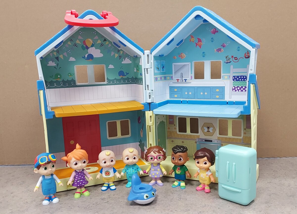 A colorful toy dollhouse with an open front shows detailed rooms. In front stand seven small toy figures, a blue toy shark, and a light blue refrigerator, all set against a plain background.
