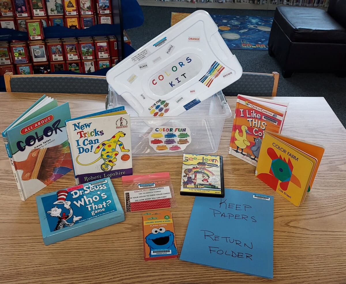 A plastic bin labeled Colors Kit contains childrens books about colors, color flashcards, a blue folder marked Keep Papers Return Folder, and crayons, all displayed on a library table.