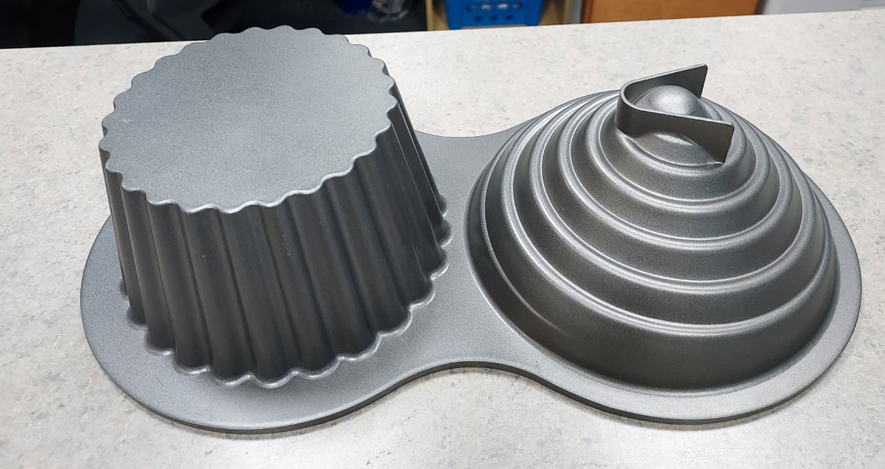A two-sided metallic baking mold sits on a countertop; one side is fluted and cylindrical, the other is dome-shaped with concentric rings and a raised triangular handle on top.