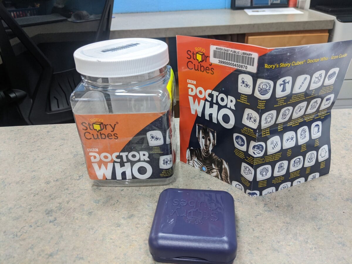 A Doctor Who-themed Rory’s Story Cubes set, including a clear plastic container, a purple case, and an unfolded instruction sheet with icons and Doctor Who branding, all on a countertop.