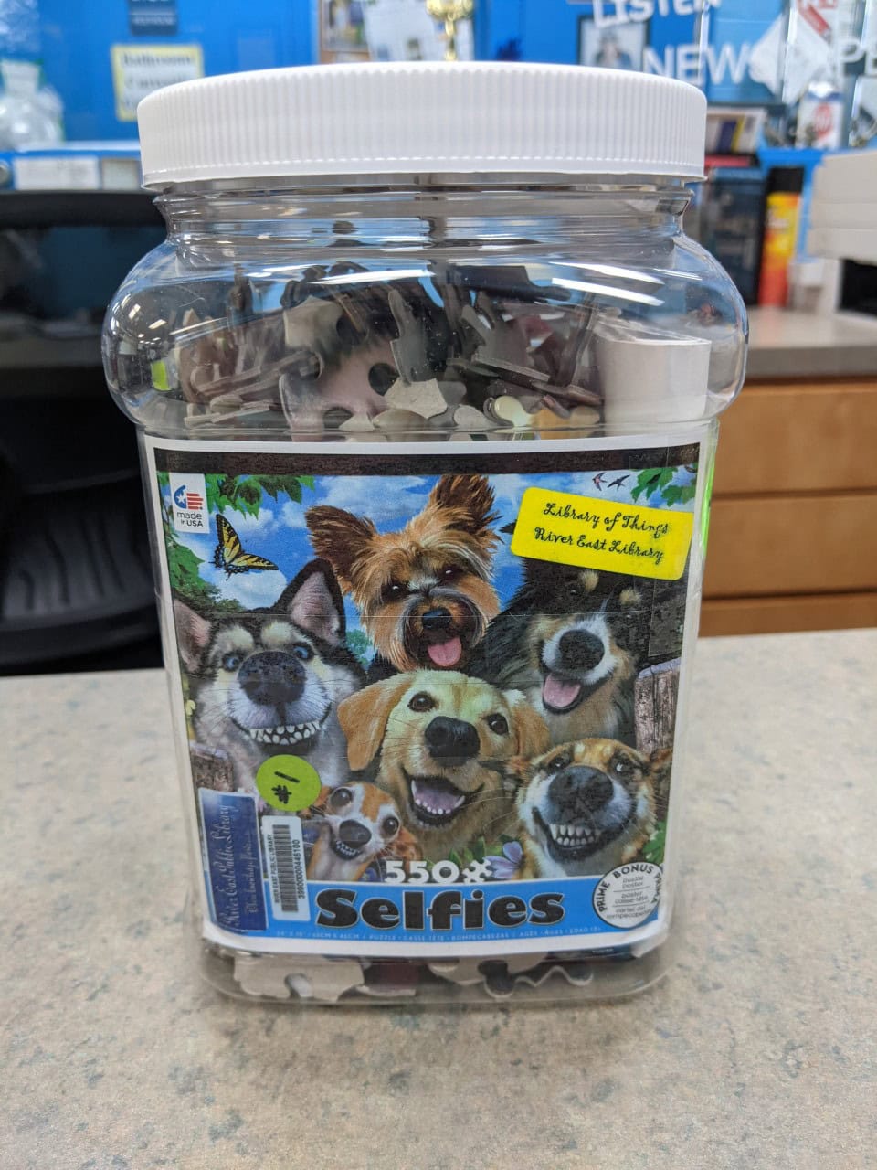 A clear plastic jar filled with puzzle pieces sits on a counter. The label shows cartoon dogs taking a selfie and reads 550 Selfies. The jar is in a library setting with books and a sign visible in the background.