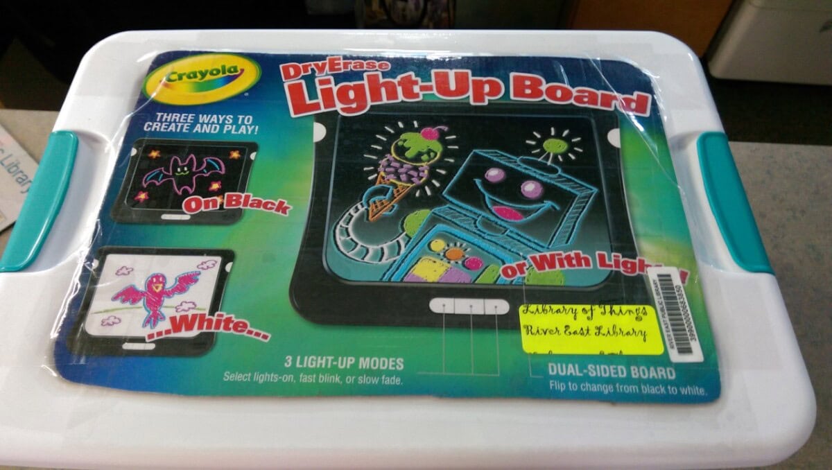 A Crayola Dry Erase Light-Up Board in its packaging, showing colorful drawings on both black and white boards with options for light-up effects. A yellow library label is also visible on the package.