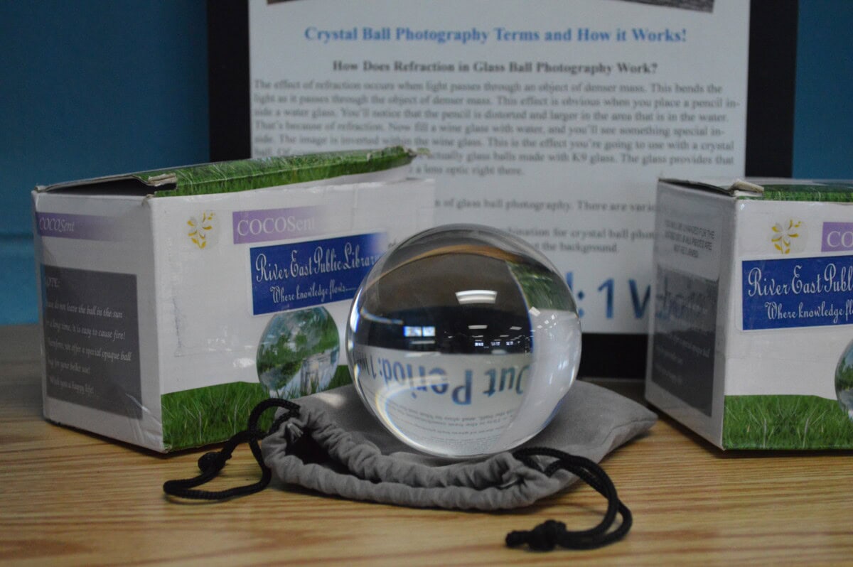A clear crystal ball sits on a gray pouch between two product boxes on a wooden surface, with an informational sign about crystal ball photography in the background. The boxes and sign are partially reflected in the crystal ball.