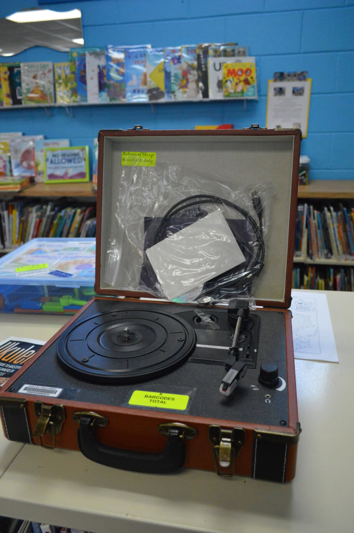 An open vintage record player sits on a library counter, with cables and a white card inside its case. Bookshelves filled with colorful childrens books are visible in the background.