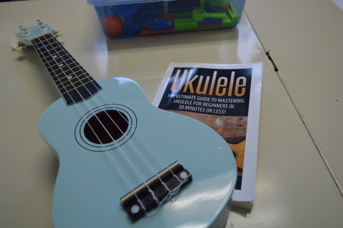 A light blue ukulele rests next to a book titled Ukulele: The Ultimate Guide to Mastering Ukulele for Beginners in 30 Minutes or Less! on a beige table, with a plastic container of colorful objects in the background.
