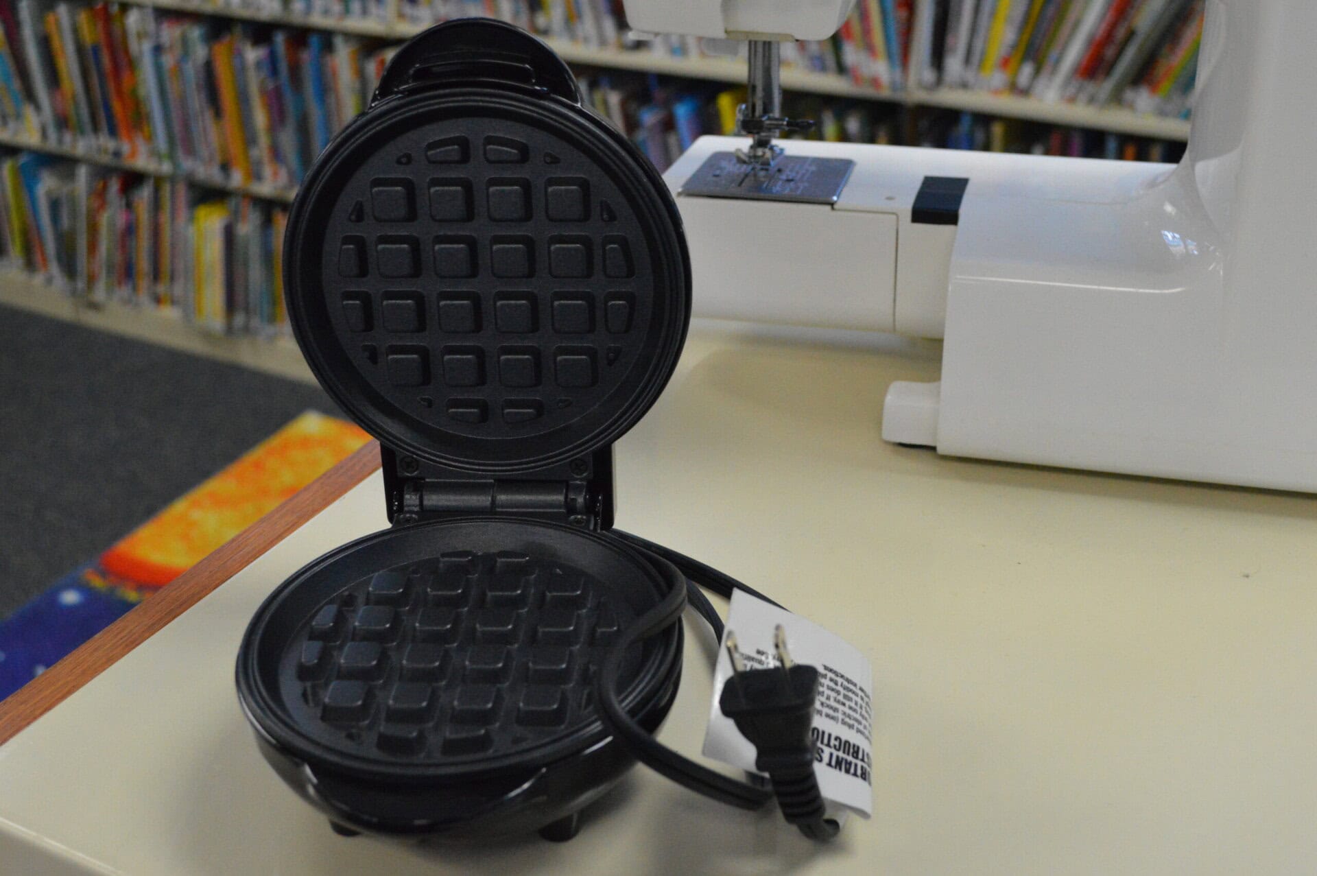 A black electric waffle maker sits open on a countertop, showing its grid plates. The power cord and plug are visible, along with a white sewing machine and bookshelves in the background.