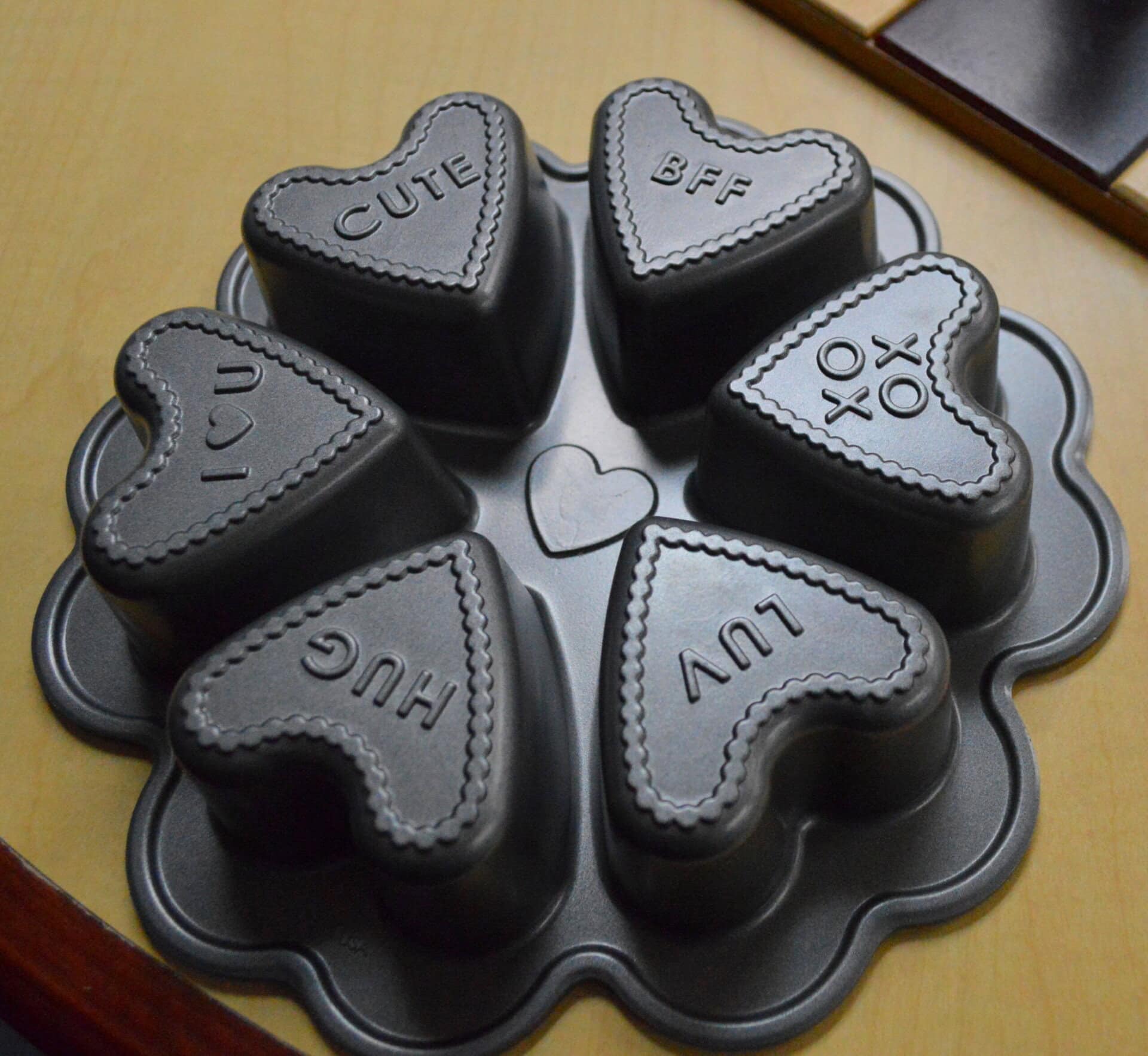 A metal baking pan with six heart-shaped molds, each labeled with a word: “CUTE,” “BFF,” “XOXO,” “LUV,” “HUG,” and “I♥U.” The pan is resting on a wooden surface.