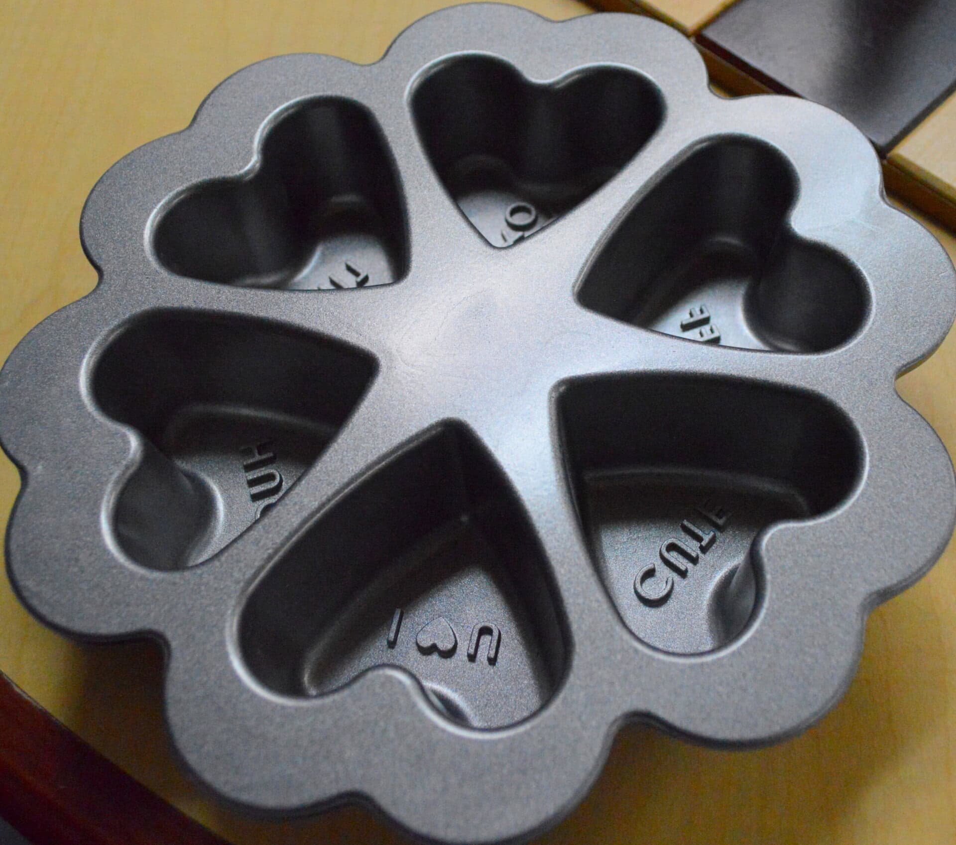 A dark gray metal baking pan with seven heart-shaped molds, each with a different message such as “I ♥ U” and “CUTIE.” The pan is placed on a light-colored wooden surface.