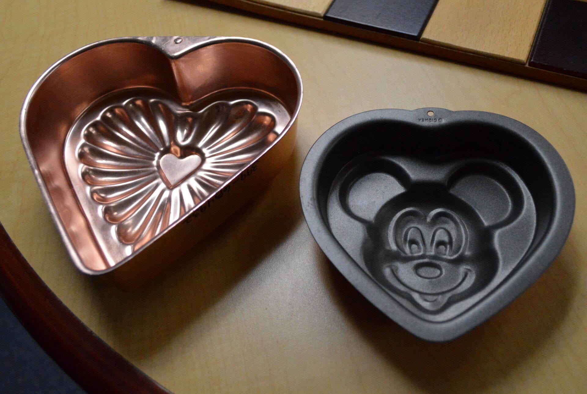 Two heart-shaped baking molds sit on a table; one is copper with a flower and heart design, and the other is dark metal with a cartoon mouse face, resembling Mickey Mouse, embossed inside.
