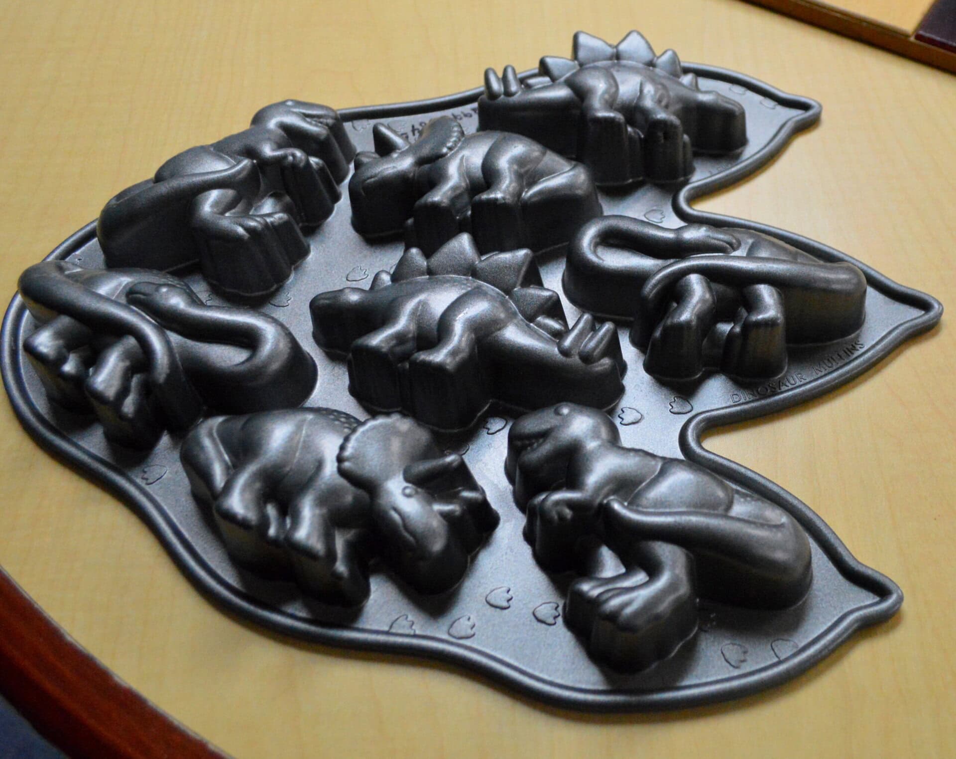 A dinosaur-shaped baking pan with molds for six different dinosaur figures, placed on a light wooden table.