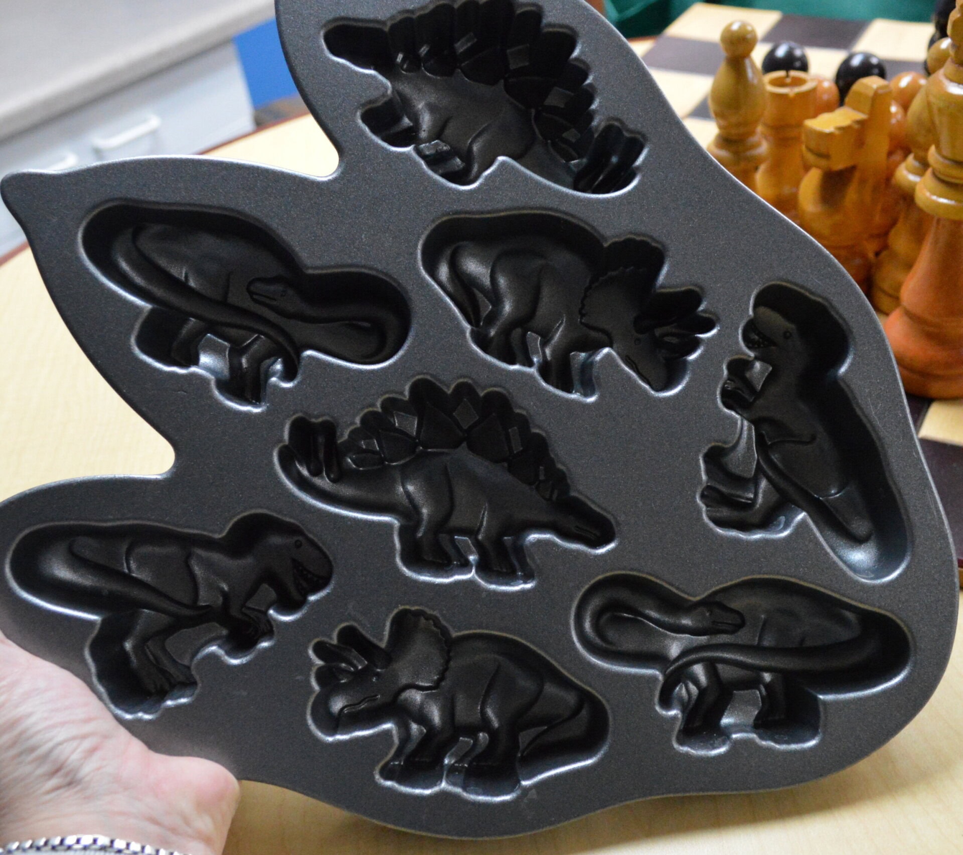 A hand holds a dark metal baking tray with seven dinosaur-shaped molds. In the background, large chess pieces are on a chessboard.