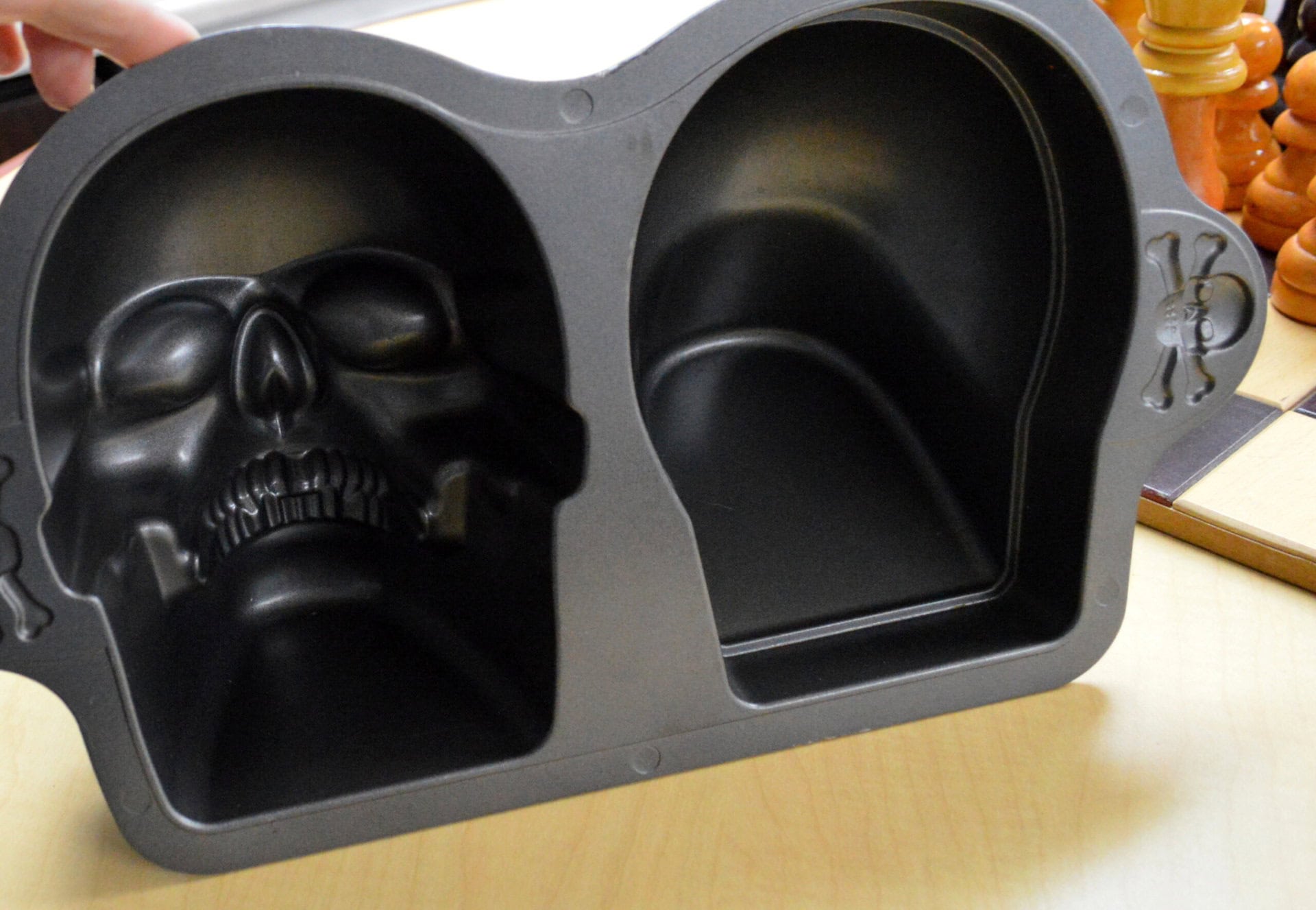 A close-up of a black skull-shaped baking pan with two compartments, one molded like a human skull and the other a plain oval, placed on a light wood surface next to a chessboard and chess pieces.