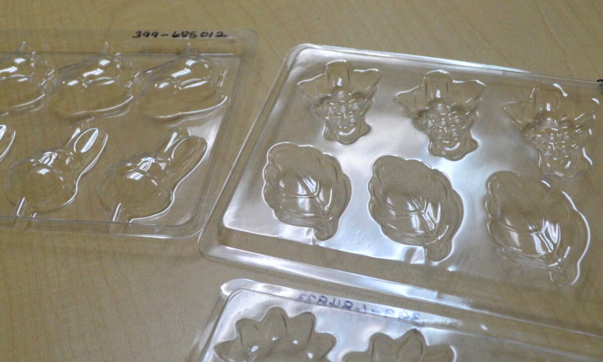 Two clear plastic molds sit on a light wood surface. The molds feature shapes resembling cartoon animal faces and leaves, intended for making chocolates or candies. Handwritten numbers appear on the edges of the molds.