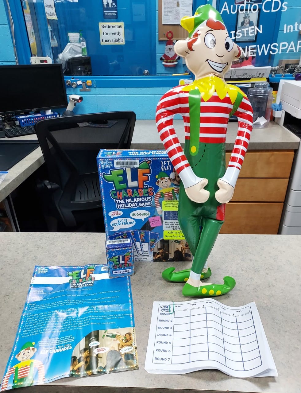 A colorful elf statue stands on a counter next to the ELF Charades game box, game sheet, and instruction booklet, with an office chair, desk, and signs in the background.
