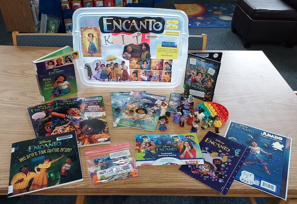 A library table displays an Encanto Kit box, several Encanto-themed books, an activity booklet, figurines of characters, a rainbow toy, and coloring materials, all related to Disney’s Encanto movie.