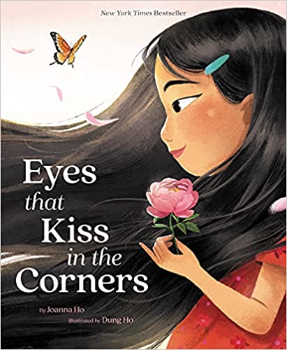 Book cover of Eyes that Kiss in the Corners by Joanna Ho. It shows a girl with long dark hair holding a pink flower, looking at a butterfly, with pink petals floating around her. Illustrated by Dung Ho.