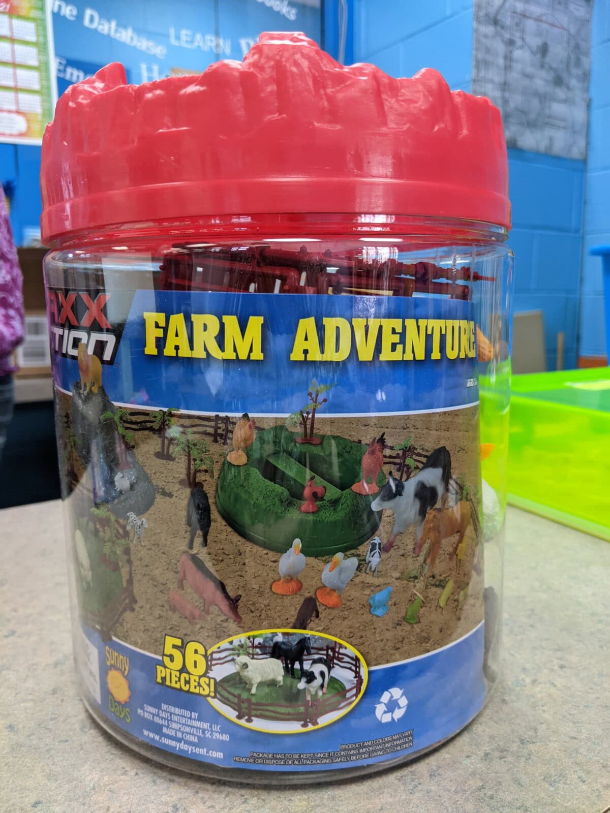 A clear plastic container with a red lid labeled Farm Adventure, showing toy farm animals and a farm scene on the label. The set contains 56 pieces and is placed on a table in a colorful room.