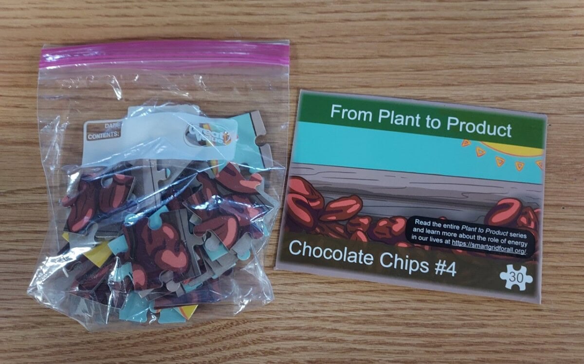 A resealable plastic bag filled with jigsaw puzzle pieces featuring cocoa beans sits next to a card titled From Plant to Product: Chocolate Chips #4 on a wood surface.