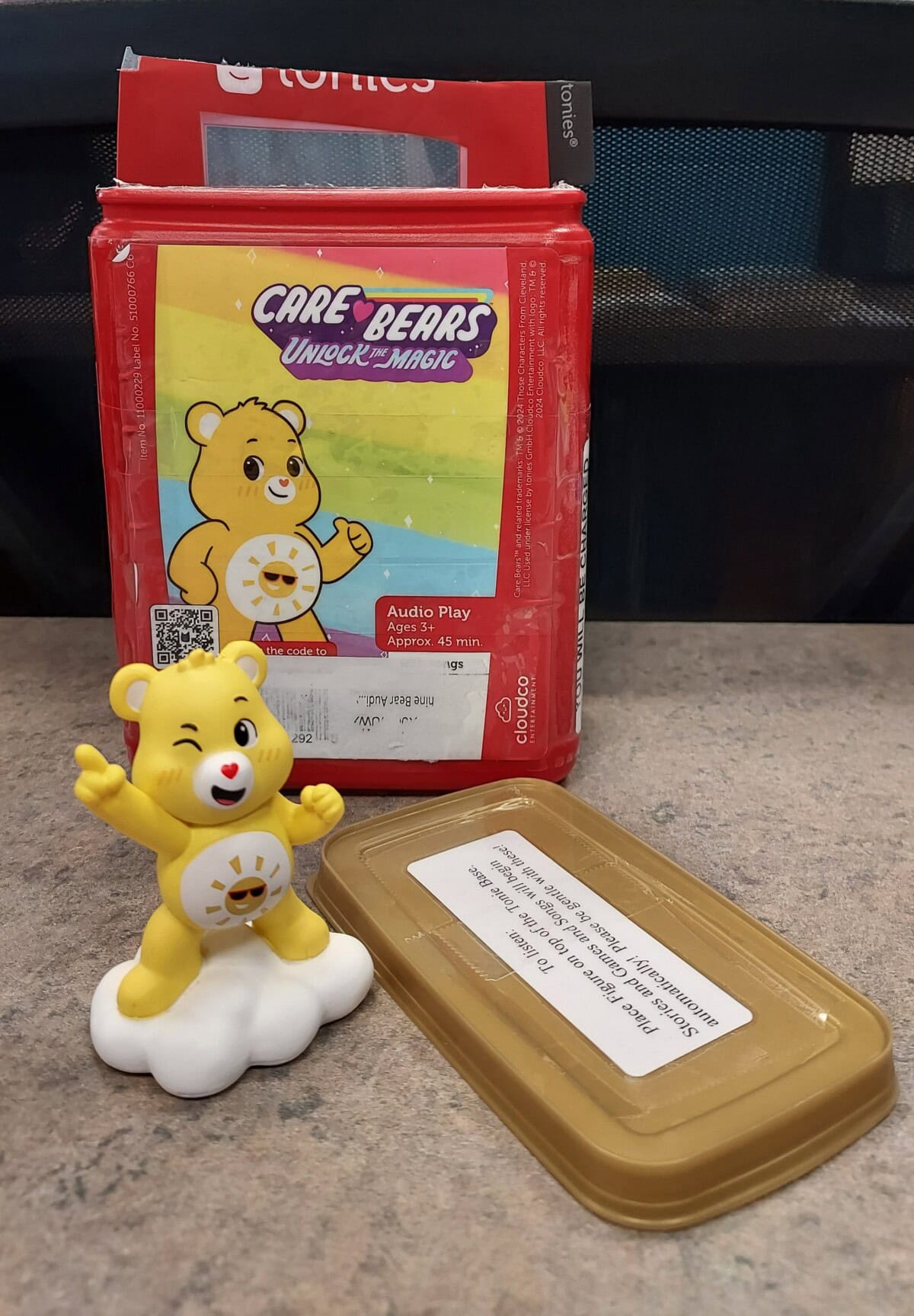 A yellow Care Bears figurine stands on a cloud, winking and waving. Behind it is a red package with Care Bears artwork and the words Unlock the Magic. A tan plastic object with a label is nearby on a gray surface.