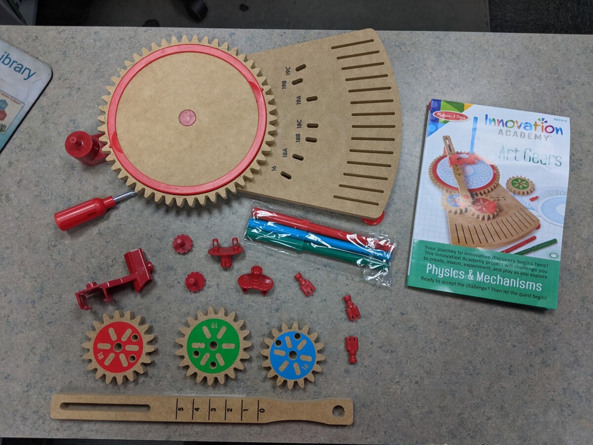 A tabletop displays a wooden gears kit with various gears, tools, and parts laid out, plus an instruction booklet titled Innovation Academy: Art Gears by Melissa & Doug.