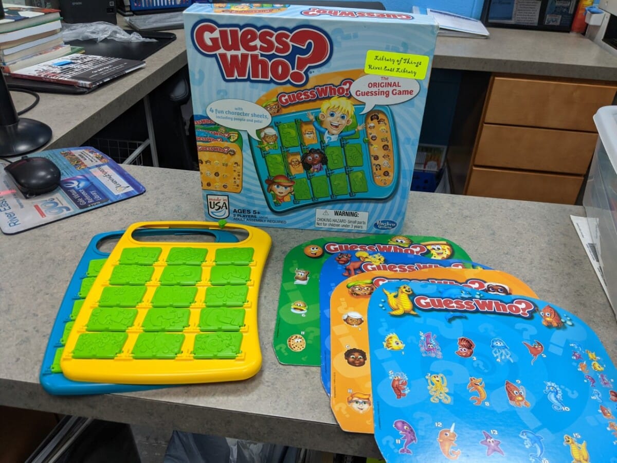 A Guess Who? board game set is on a countertop, featuring the game box, two plastic game boards, and several character sheets with colorful illustrations and cartoon faces.
