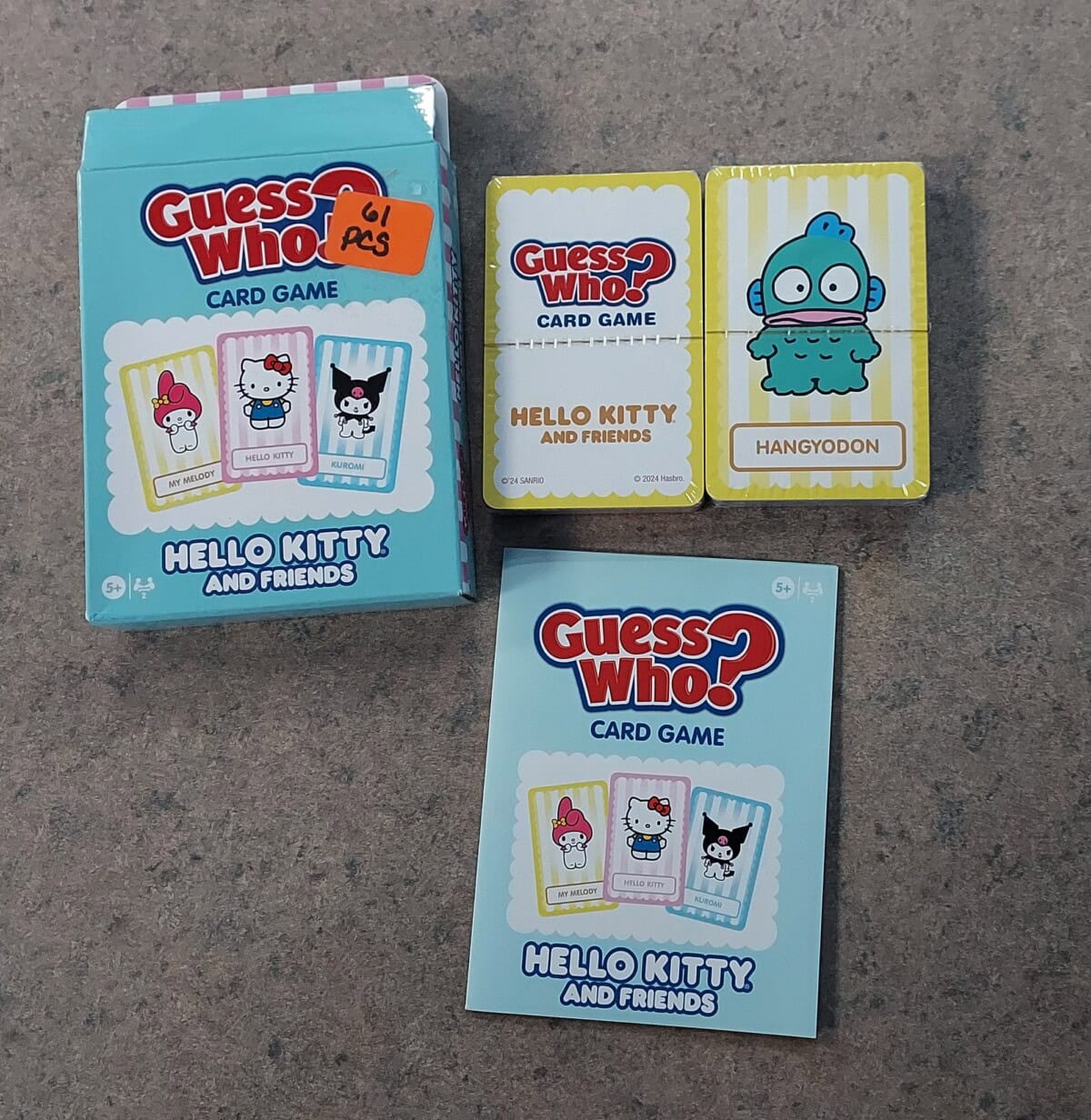 A Guess Who? Hello Kitty and Friends card game is shown. The box, a rule card, and cards featuring Hello Kitty, My Melody, Kuromi, and Hangyodon characters are laid out on a gray surface.