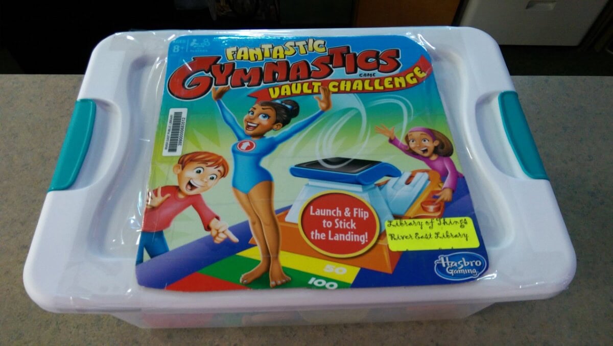 Board game box for Fantastic Gymnastics Vault Challenge by Hasbro Gaming, featuring cartoon gymnasts on the cover and text promoting flipping and sticking the landing. The box rests on a white plastic container.