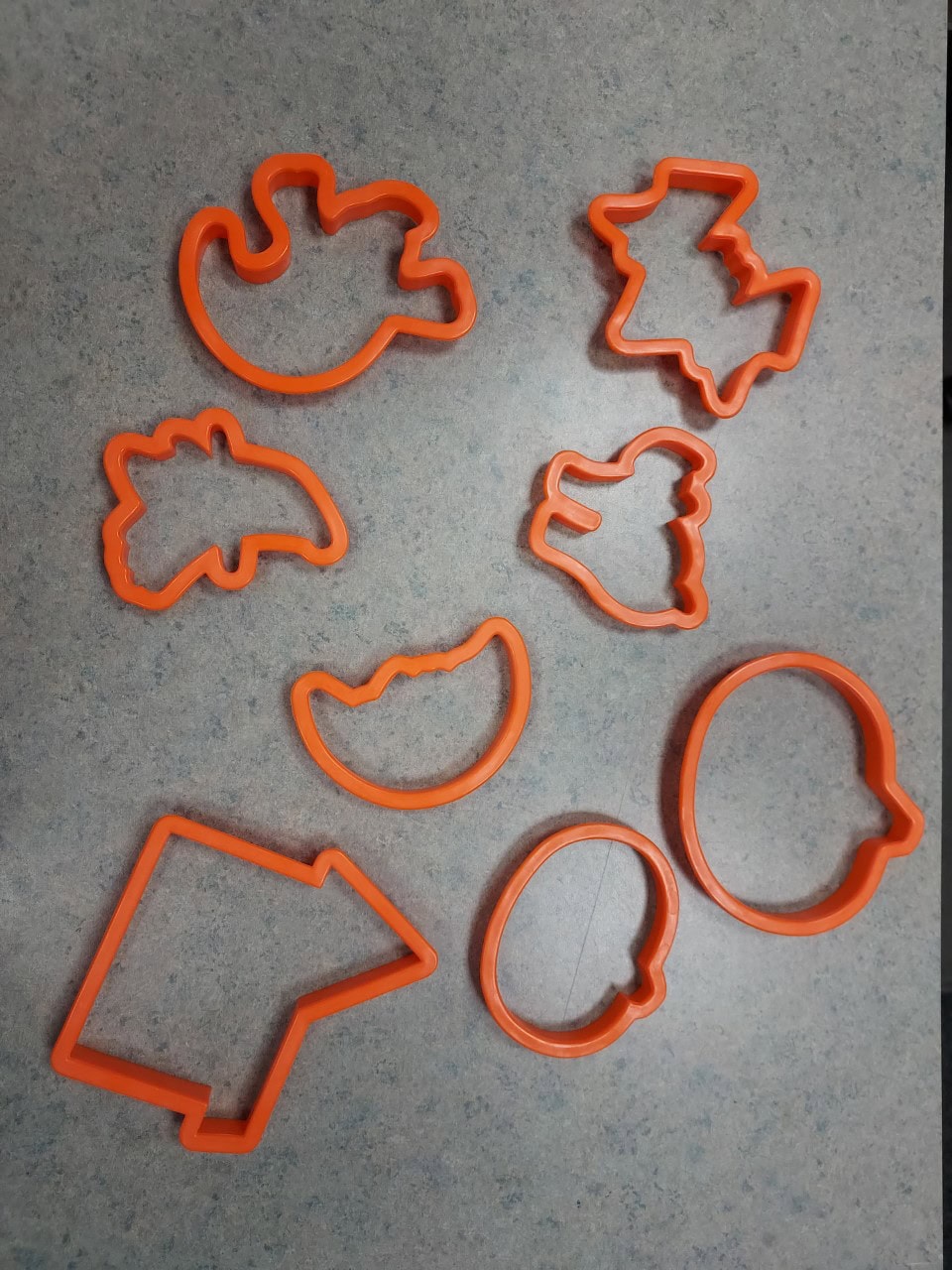 Eight orange plastic cookie cutters in various shapes, including a pumpkin, ghost, bat, witch hat, cat, crescent moon, arrow, and oval, are arranged on a gray speckled surface.