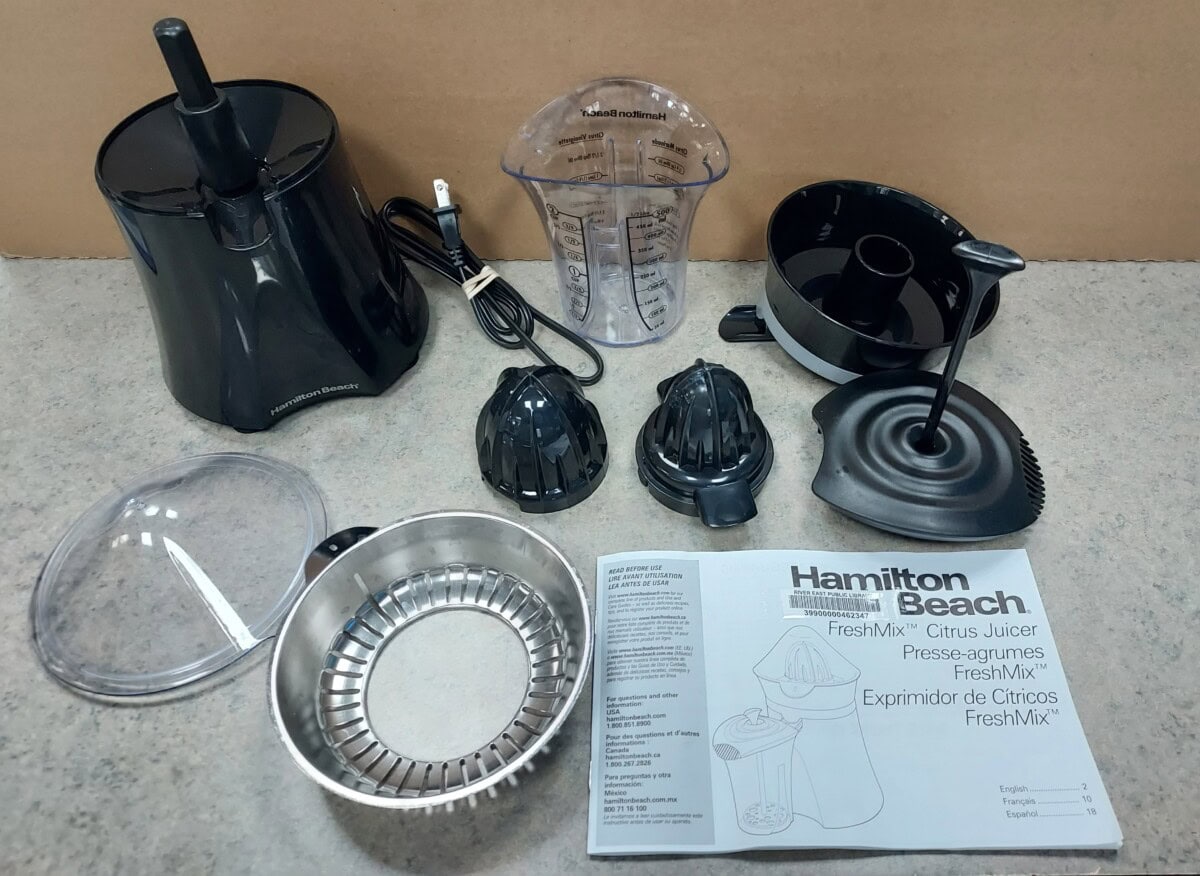 A disassembled Hamilton Beach citrus juicer is shown with all its parts, including the base, juicing cone, pulp collector, measuring cup, lid, reamers, and instruction manual, displayed on a countertop.